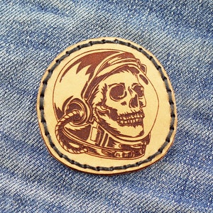 May include: A brown leather patch with a stitched border, featuring a detailed illustration of a skull wearing an astronaut helmet.