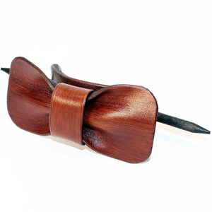 May include: A brown leather bow tie hair clip with a black metal pin. The bow tie is made of a soft, supple leather and has a classic, elegant design.