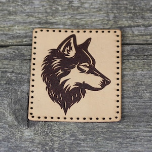 May include: A brown leather patch with a black silhouette of a wolf's head. The patch has a stitched border.