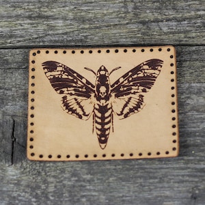 Könnte beinhalten: Ein braunes Lederstück mit einem schwarzen und weißen Motten-Design. Die Motte hat ein Totenkopf- und Knochenkreuzmuster auf ihrem Körper.