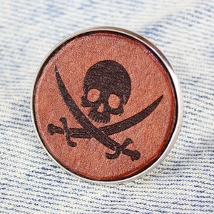 Peut inclure: Une épingle en cuir marron avec un dos argenté représentant un crâne et des os croisés noirs.