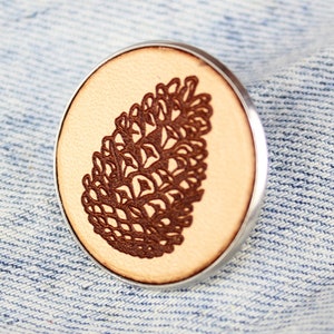 May include: A wooden pin with a silver backing featuring a detailed brown pine cone design.