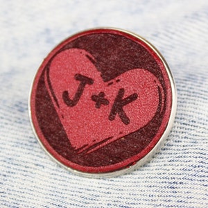 Może przedstawiać: Czerwona przypinka w kształcie serca z srebrną obwódką. Serce ma napisane w środku litery "J*K".