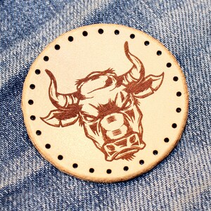Puede incluir: Una pieza redonda de cuero de color beige claro con un grabado marrón detallado de la cabeza de un toro. El toro tiene cuernos grandes y una expresión intensa. El borde del cuero tiene agujeros de costura negros espaciados uniformemente. El fondo es tela vaquera azul.