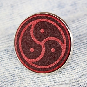 Könnte beinhalten: Eine rote und silberne Anstecknadel mit einem Symbol aus drei ineinandergreifenden Kreisen. Die Anstecknadel befindet sich auf einem blauen Jeansstoff.