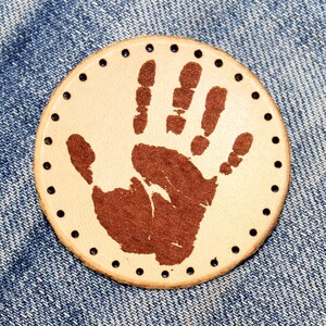 May include: A round, beige leather patch with a brown handprint in the center. The patch has a circle of evenly spaced holes around the edge.