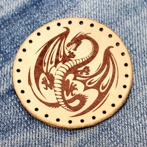 May include: A round, beige leather patch with a brown dragon design. The dragon is curled up with its wings spread out. The patch has a circle of small holes around the edge.