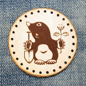 May include: A brown leather patch with a cartoon image of a mole holding a shovel and a small plant. The patch has a circle of holes around the edge.