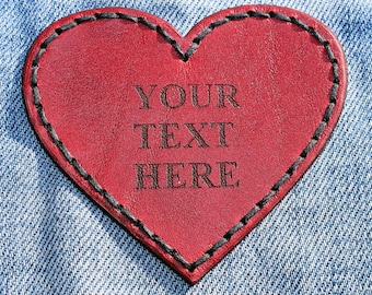 Puede incluir: Un parche de cuero rojo en forma de corazón con costuras negras alrededor de los bordes. El texto "YOUR TEXT HERE" está impreso en negro en el parche.
