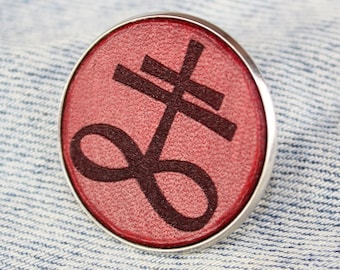 Könnte beinhalten: Eine runde, silberne Anstecknadel mit rotem Hintergrund und einem schwarzen Symbol. Das Symbol ist ein stilisiertes Kreuz mit einer Schleife am unteren Ende.