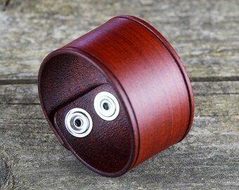 Puede incluir: Un brazalete ancho de cuero marrón oscuro con dos cierres de botón a presión plateados. El brazalete es una banda sólida de cuero, con una forma ligeramente curvada. El brazalete se muestra sobre una superficie de madera envejecida.