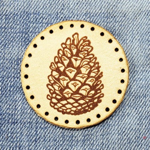 Könnte beinhalten: Ein braunes Lederstück mit einer detaillierten Illustration einer Kiefernzapfen in der Mitte. Das Stück hat einen Kreis aus kleinen Löchern am Rand.