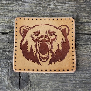 Könnte beinhalten: Ein braunes Lederpatch mit einem braunen Bärenkopfdesign. Der Bär brüllt mit offenem Maul und sichtbaren Zähnen. Das Patch hat einen genähten Rand.