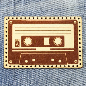 以下が含まれることがあります： 黄色の背景に茶色の革でできた、ヴィンテージスタイルのカセットテープのデザイン。点線の縁取りで縫製されています。カセットには「Side A」の文字が印刷されています。