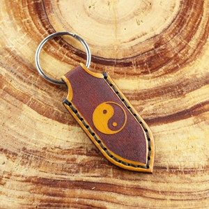 Könnte beinhalten: Ein brauner Leder-Schlüsselanhänger mit einem gelben Yin-Yang-Symbol. Der Schlüsselanhänger hat einen silbernen Schlüsselring.