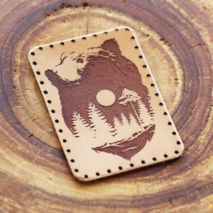 May include: A brown leather patch with a brown bear silhouette and a forest scene with a full moon. The patch has a stitched border.