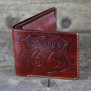 Puede incluir: Una cartera de cuero marr&oacute;n con un borde cosido verde. La cartera tiene un dise&ntilde;o de la Ruta 66 grabado en relieve en la parte delantera.