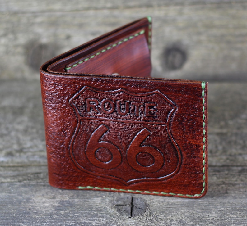 Puede incluir: Una cartera de cuero marr&oacute;n con un borde cosido verde. La cartera tiene un dise&ntilde;o de la Ruta 66 grabado en relieve en la parte delantera.