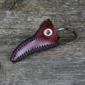 May include: A brown leather keychain shaped like a bird's beak with a silver metal ring. The keychain has black stitching around the edges.