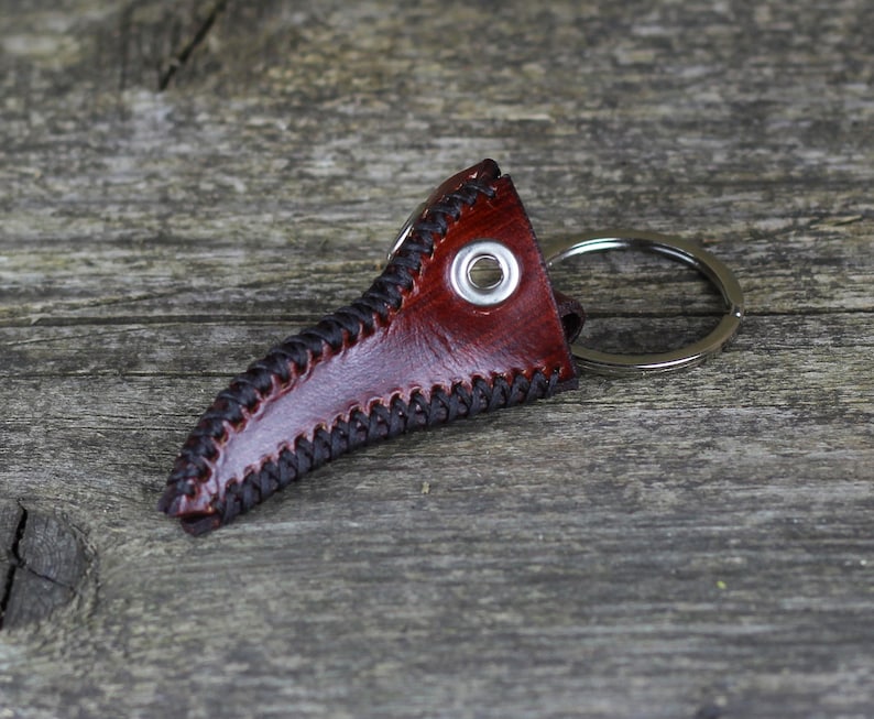 May include: A brown leather keychain shaped like a bird's beak with a silver metal ring. The keychain has black stitching around the edges.