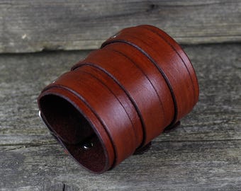 May include: A wide, brown leather cuff bracelet with multiple straps and a buckle closure. The cuff is made of a rich, reddish-brown leather and appears to be handcrafted. The bracelet is designed to be worn around the wrist.