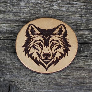 Könnte beinhalten: Ein brauner Lederuntersetzer mit einem schwarzen Wolfskopfdesign. Die Augen des Wolfes sind geschlossen und sein Maul ist leicht geöffnet.