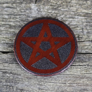 Può includere: Un sottobicchiere rotondo in legno con un design a pentagramma rosso su sfondo nero.