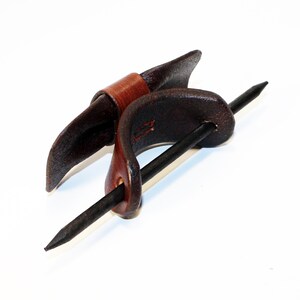 May include: A brown leather hair stick with a dark brown leather wrap. The stick is made of metal and has a pointed end.