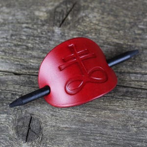 May include: A red leather hair stick with a black wooden pin. The leather has a raised design of a cross and an infinity symbol.