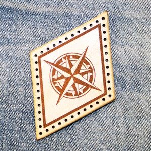 May include: A brown leather patch with a white compass design. The compass has four points and a star in the center. The patch has a decorative border of small holes.