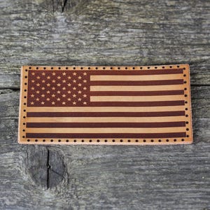 Puede incluir: Un parche rectangular de cuero con el diseño de la bandera estadounidense. La bandera tiene un campo de estrellas y rayas rojas y blancas alternas. El parche es de color marrón claro con detalles en marrón oscuro y un borde de pequeños agujeros.