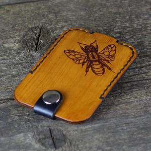 May include: A small, light brown leather card holder with a black stitched border and a bee design. The card holder has a black strap with a silver metal button.