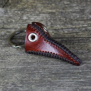 May include: A brown leather plague doctor mask keychain with black stitching and a silver key ring.