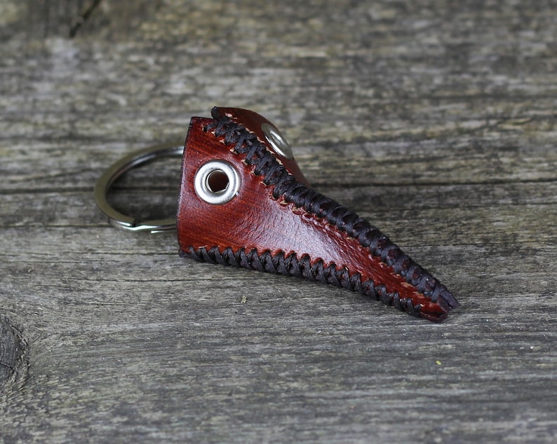 May include: A brown leather plague doctor mask keychain with black stitching and a silver key ring.
