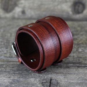 Op de afbeelding: Een bruin leren armband met een zilveren gesp. De armband is gemaakt van twee lagen leer, waarbij de bovenste laag over de onderste laag is gevouwen.