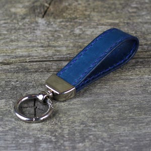 Puede incluir: Un llavero de cuero azul con un anillo de metal plateado y un lazo de metal plateado. El llavero está cosido con hilo blanco.