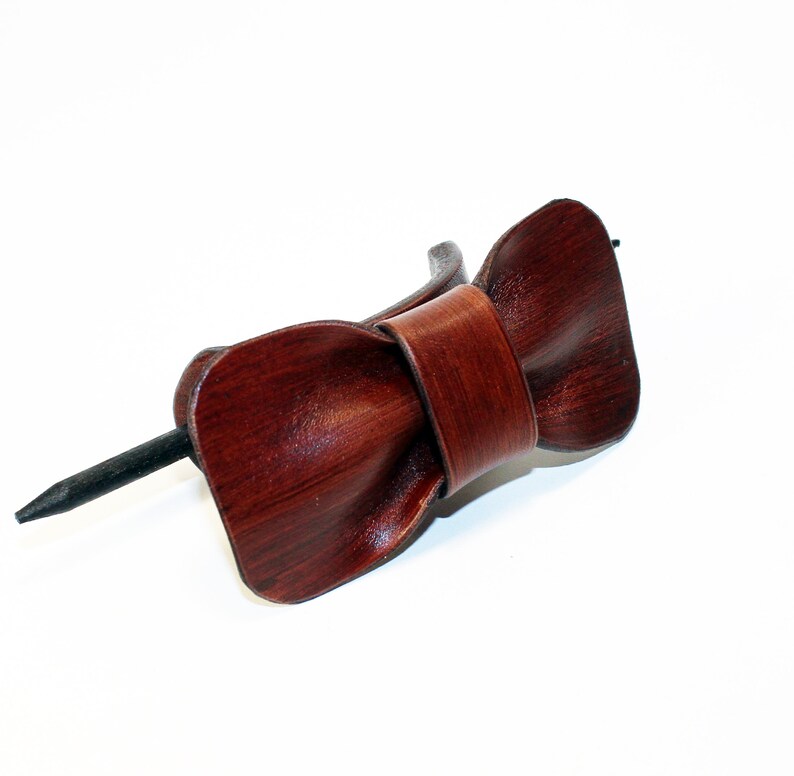 May include: A brown leather hair stick with a bow shape. The stick is made of metal and is black.