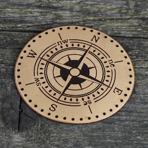 Könnte beinhalten: Ein runder, brauner Lederkompass mit einer schwarzen Kompassrose und den Richtungen N, S, E, W, NE, NW, SE und SW, die darauf eingraviert sind.