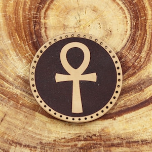 May include: A brown leather coaster with a gold ankh symbol in the center. The coaster has a decorative stitched edge.