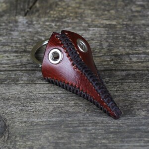 May include: A brown leather plague doctor mask keychain with black stitching and a silver metal ring.