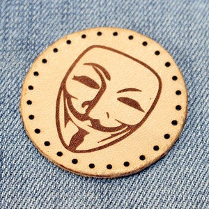 Könnte beinhalten: Ein runder, brauner Lederfleck mit schwarzem Rand und einem schwarz-weißen Bild der Guy-Fawkes-Maske. Die Maske hat ein breites Lächeln und eine spitze Nase.