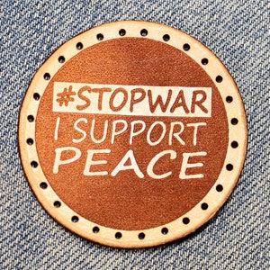 May include: A brown leather patch with white stitching and the text "#STOPWAR I SUPPORT PEACE" in white lettering.