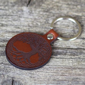 Peut inclure: Un porte-clés rond en cuir marron avec un motif détaillé d'arbre de vie. Il est doté d'un anneau en métal argenté et d'une petite boucle en cuir. Le motif de l'arbre est en relief sur le cuir, créant un effet texturé.