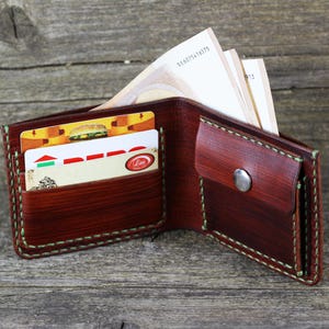 Puede incluir: Una cartera de cuero marr&oacute;n con una solapa y un cierre de presi&oacute;n. La cartera est&aacute; abierta y tiene varias tarjetas y billetes dentro. La cartera tiene un detalle de costura verde.