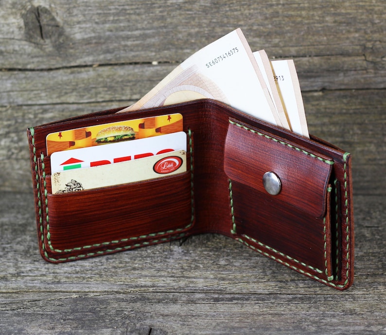 Puede incluir: Una cartera de cuero marr&oacute;n con una solapa y un cierre de presi&oacute;n. La cartera est&aacute; abierta y tiene varias tarjetas y billetes dentro. La cartera tiene un detalle de costura verde.