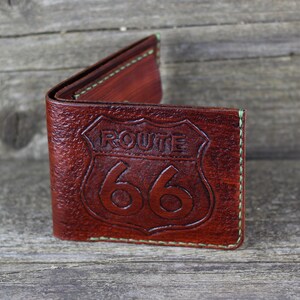 Puede incluir: Una cartera de cuero marr&oacute;n con un dise&ntilde;o de la Ruta 66 en relieve en la parte delantera. La cartera tiene costuras verdes alrededor de los bordes.