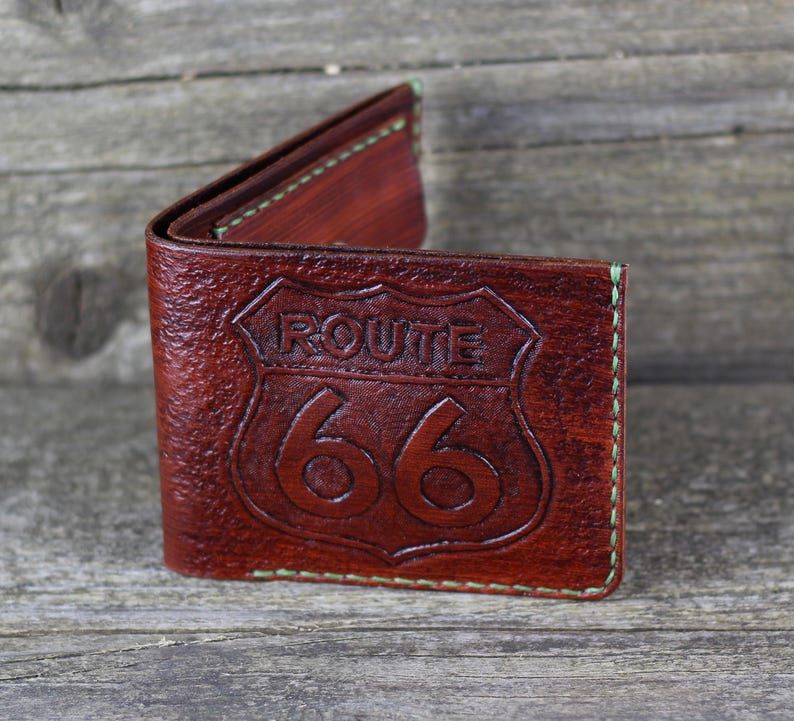 Puede incluir: Una cartera de cuero marr&oacute;n con un dise&ntilde;o de la Ruta 66 en relieve en la parte delantera. La cartera tiene costuras verdes alrededor de los bordes.