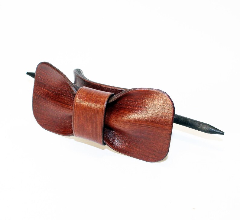 May include: A brown leather bow tie hair clip with a black metal pin. The bow tie is made of a soft, supple leather and has a classic, elegant design.