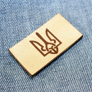 May include: A brown leather patch with a brown embroidered Ukrainian trident symbol.