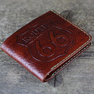 Puede incluir: Una cartera de cuero marr&oacute;n con un borde cosido verde. La cartera presenta un dise&ntilde;o de escudo Route 66 en relieve.